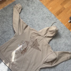 Beige hoodie från Jordan x Travis Scott - Säljer en beige hoodie från Jordan x Travis Scott med broderad design på bröstet och en stor logga på ryggen. Hoodien har en känguruficka och långa ärmar med en röd logga på ärmslutet. Perfekt för streetwear-stilen! Hör av er vid flera funderingar!!