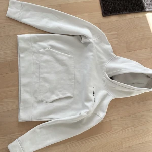 Vit hoodie med fic - Säljer en stilren vit hoodie med en praktisk magficka och en liten textdetalj på bröstet. Perfekt för en avslappnad look. Hoodien har långa ärmar och en bekväm passform.