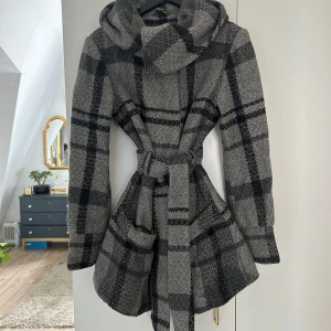 Grå och svart rutig kappa - Snygg grå och svart rutig kappa med knappar och bälte i midjan. Den har en stor krage och långa ärmar, perfekt för kyligare dagar. Kappan har ett klassiskt mönster och är både stilren och bekväm.