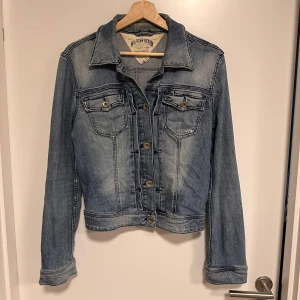 Blå jeansjacka från Hilfiger Denim - Snygg blå jeansjacka från Hilfiger Denim med klassisk design och knappar framtill. Jackan har två bröstfickor med knapp och långa ärmar. Lite kortare modell. Lappen i nacken något missfärgad. Perfekt för en avslappnad stil.