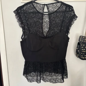 Svart spetsblus med korsett - Elegant svart blus med spetsdetaljer och inbyggd korsett. Blusen har en vacker öppen rygg och dragkedja på sidan. Perfekt för en stilfull look. Storlek M men passar bättre en S!❤️