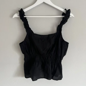 Svart linnetopp med volangdetaljer - Snygg svart linnetopp med volangdetaljer på axelbanden. Toppen har en elastisk midja som ger en smickrande passform. Perfekt för varma sommardagar eller en kväll ute.