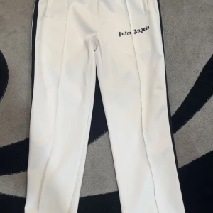 Vita byxor från Palm Angels - Snygga vita byxor från Palm Angels med svarta sidopaneler och logotyp på framsidan. Byxorna har en normal passform och är perfekta för en stilren look. De är tillverkade i ett bekvämt material och har en sportig känsla.