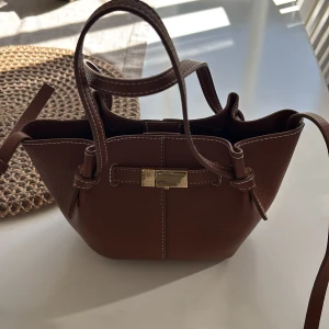 Brun axelväska med gulddetalj - Snygg brun axelväska i skinn med en elegant gulddetalj på framsidan. Väskan har både handtag och en justerbar axelrem för flexibilitet. HELT SLUTSÅLD därav priset