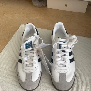 Adidas Samba sneakers - Snygga Adidas Samba sneakers i vitt med svarta detaljer och klassiska tre ränder. Skorna har en stilren design med snörning och en brun sula. Helt nya, aldrig använda 