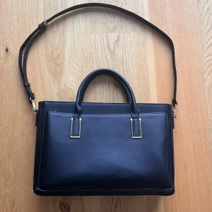 Svart axelväska med handtag - Snygg svart axelväska i skinn med både handtag och justerbar axelrem. Väskan har en stilren design och är perfekt för att bära dina dagliga nödvändigheter. Insidan är rymlig med flera fack för enkel organisering. Ytterfacket är perfekt för att lägga dator och data fodral i.