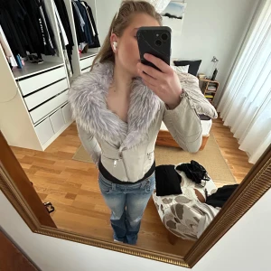 Beige jacka med fuskpäls från River Island - Snygg beige jacka från River Island med en stor krage i grå fuskpäls. Jackan har dragkedja framtill och två dragkedjefickor. Perfekt för kyligare dagar med sin stilrena design och bekväma passform.