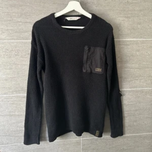 Svart stickad tröja från H&M - Säljer en svart stickad tröja från H&M med en praktisk bröstficka med dragkedja. Tröjan har en rund halsringning och långa ärmar, perfekt för kyligare dagar.