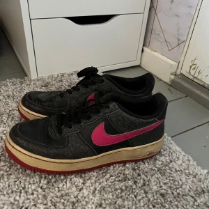 Nike Air Force med rosa detaljer - Snygga Nike Air Force sneakers i svart med rosa swoosh och detaljer. Skorna har en klassisk design med snörning och en robust sula i rött. Perfekta för en cool och avslappnad stil.