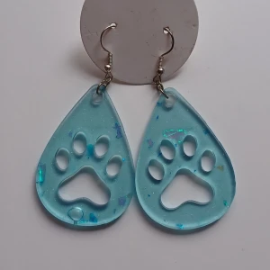 Blå örhängen med tassmotiv - Dessa är 2a utrensning! Unika droppformade örhängen i blått med utskuret tassmotiv. De har en glittrig finish och är perfekta för djurälskare. Lätta att bära med krokfäste. 