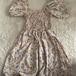 Blommig klänning 🌸 - Charmig blommig klänning med puffiga kortärmar och smockdetaljer. Klänningen har en mjuk färgpalett med inslag av blått och beige, perfekt för vår och sommar. Den har en bekväm passform och ett romantiskt utseende.🌺🌺