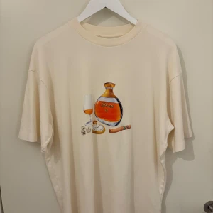 Beige t-shirt från Drôle de Monsieur - Säljer en beige t-shirt från Drôle de Monsieur med ett tryck av en flaska, glas och cigarr på framsidan. T-shirten har en avslappnad passform och korta ärmar. Perfekt för en stilren och avslappnad look. I storlek L
