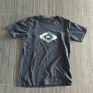 Svart t-shirt från Quiksilver - Säljer en svart t-shirt från Quiksilver med ett tryck på bröstet. T-shirten är kortärmad och gjord i bomull, perfekt för en avslappnad stil. Passar bra till både jeans och shorts. Hör av dig vid intresse! Pris kan diskuteras. 