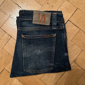 Mörkblå jeans från Nudie Jeans - Snygga mörkblå jeans från Nudie Jeans med klassisk femficksdesign och orange sömmar. Jeansen har en straight passform och är tillverkade i högkvalitativt denim. Perfekta för en stilren look.