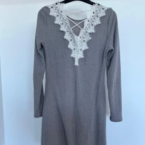 Grå ribbad klänning med spetsdetaljer - Snygg grå ribbad klänning med vacker vit spets och snörning vid halsen. Klänningen har långa ärmar och en bekväm passform. Perfekt för en stilren look.
