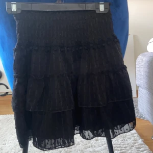 Svart kjol från Shein - Snygg svart kjol från Shein med volanger och elastisk midja. Kjolen har ett subtilt prickigt mönster och är perfekt för att ge en chic touch till din outfit. Den är kort och passar bra till olika tillfällen.