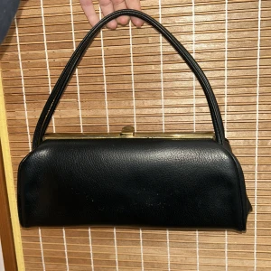 Svart handväska med metallspänne - Elegant svart handväska i skinn med ett stilrent metallspänne upptill. Väskan har dubbla handtag. Mindre defekter på insidan på grund av ålder. 