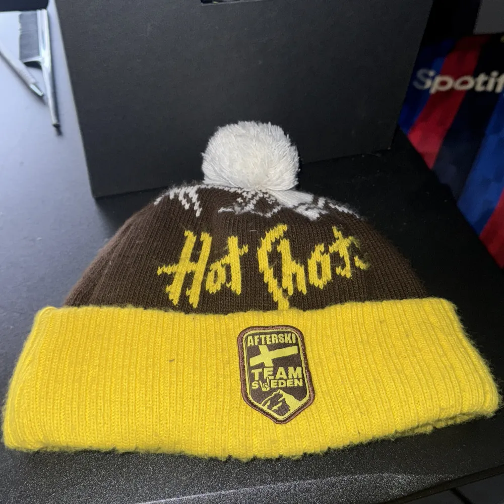 Säljer en cool brun och gul mössa med texten 'Hot shot' och ett märke med 'Afterski Team Sweden'. Mössan har en vit tofs på toppen och ett snöflingemönster.. Asusteet.