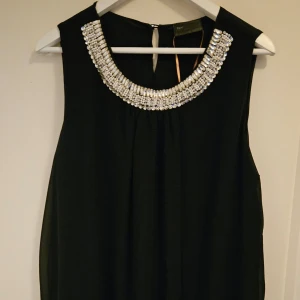 Svart ärmlös klänning med pärldetaljer - Elegant svart ärmlös klänning från bpc selection Premium. Klänningen har en vacker pärlutsmyckning runt halsringningen som ger en lyxig känsla. Perfekt för speciella tillfällen. Är ungefär 95 cm i längden