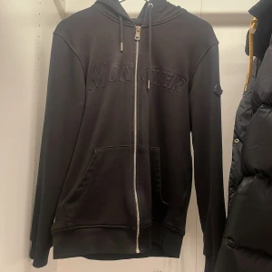 Svart hoodie från Moncler - Säljer en stilren svart hoodie från Moncler med dragkedja och broderad logga på bröstet. Jackan har långa ärmar och en praktisk huva. Perfekt för en casual look.