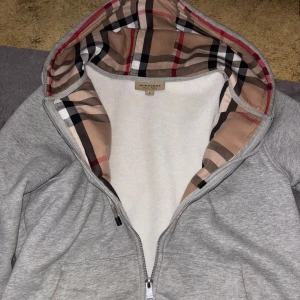 Grå hoodie från Burberry - Säljer en stilren grå hoodie från Burberry med klassiskt rutigt mönster i huvan. Den har dragkedja framtill och en broderad logga på bröstet. Perfekt för en avslappnad look. Säljer för att den inte passar mig längre