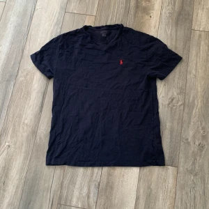 Mörkblå t-shirt från Ralph Lauren - Säljer en stilren mörkblå t-shirt från Ralph Lauren med en röd broderad logga på bröstet. Perfekt för en avslappnad look. T-shirten har korta ärmar och är tillverkad i mjukt bomullsmaterial. Tröjan har en liten fläck som går att tvätta bort, jag skulle säga att storleken är S men passar M
