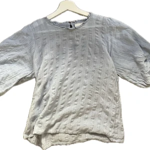 Ljusblå blus med struktur - Säljer en ljusblå blus med ett subtilt randigt mönster och struktur. Blusen har trekvartsärmar och en knappdetalj på ryggen. Perfekt för sommaren