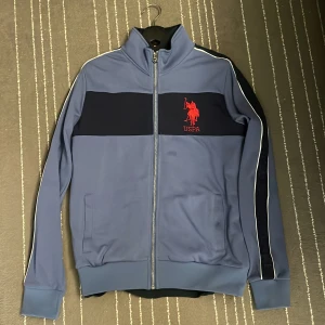 Blå tröja från U.S. Polo Assn. - Säljer en snygg och skön blå tröja från U.S. Polo Assn. med dragkedja och röd logo på bröstet. Tröjan har långa ärmar med vita & mörkblå detaljer längs sidorna och är tillverkad i en bomulls- och polyesterblandning (60/40).