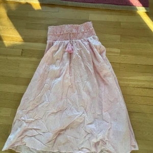 Rosa långkjol från Gina Tricot - Säljer en söt rosa långkjol från Gina Tricot med smockdetaljer i midjan och ett diskret mönster. Kjolen har en avslappnad passform och är perfekt för varma dagar. Den är lätt och luftig, vilket gör den bekväm att bära hela dagen. Passar perfekt med en enkel topp eller blus.