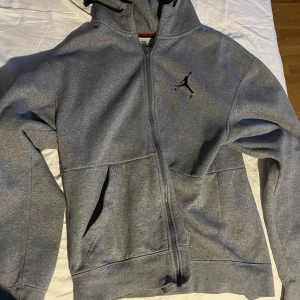 Grå hoodie från Jordan - Säljer en grå hoodie från Jordan med dragkedja och det klassiska logotyptrycket på bröstet. Tröjan har en justerbar huva och praktiska fickor framtill. Perfekt för en avslappnad stil.
