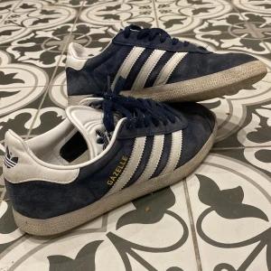 Adidas Gazelle i blå mocka - Säljer ett par klassiska Adidas Gazelle sneakers i blå mocka med vita detaljer och snörning. Skorna har den ikoniska tre-randiga designen och en bekväm sula. Perfekta för en stilren och sportig look.