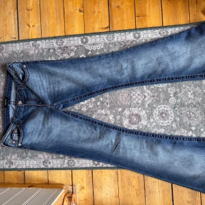 Blå jeans från True Religion - Snygga blå jeans från True Religion x NELLY med kontrasterande röd söm och klassisk femficksdesign. Jeansen har en bootcut passform och är i perfekt sick. Märkets ikoniska broderade fickor ger en extra touch. Är i strlk 31 , säljer pga passar inte och har inte kommit till användning plagget användes en gång och tvättades en gång annars har dem inte använts, pris går att förhandla kontakta bara🤗