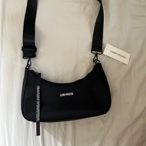Svart axelväska från Carin Wester - Snygg svart axelväska från Chuck Hester med justerbar rem och dragkedja. Perfekt för att bära dina nödvändigheter med stil. Väskan har en minimalistisk design med märkets logga framtill och på dragkedjan. Prislappen är kvar. 