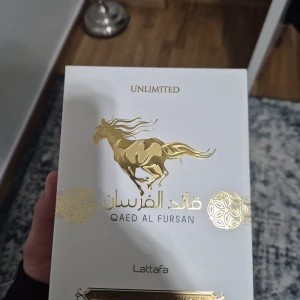 Qaed Al Fursan Parfym - Elegant parfym från Lattafa, Qaed Al Fursan Unlimited, i en stilren vit flaska med gyllene detaljer och ett unikt mönster. Flaskan har en gyllene kork och en design som utstrålar lyx och elegans. Perfekt för den som söker en sofistikerad doftupplevelse. Helt ny men inte nöjd men doften jag är mer åt de mörkare hållet