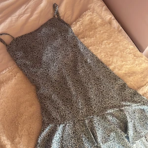 Blommig blå klänning med volang - En blå klänning med ett sött blommönster och volangdetaljer och med en rosett i ryggen som går att knyta hur man vill. Klänningen har smala axelband och som går att spänna in och ut.