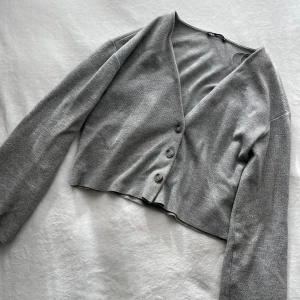 Grå kofta med knappar - Säljer en stilren grå kofta med tre knappar framtill. Koftan har en v-ringad design och är tillverkad i ett mjukt material. Perfekt för lager-på-lager under kyligare dagar. Koftan är kort som passar till vår och sommar