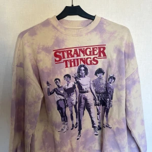 Lila Stranger Things tröja från H&M - Säljer en cool lila batikmönstrad tröja från H&M med Stranger Things-tryck på framsidan. Tröjan har långa ärmar och en rund halsringning. Perfekt för fans av serien!