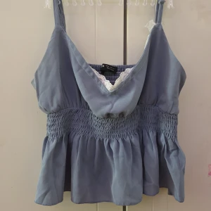 Blå linnetopp med spetsdetaljer - Snygg blå linnetopp med smockdetaljer och spetskant vid halsringningen som jag själv har sytt dit. Toppen har en lös passform och är perfekt för varma dagar. Den har breda axelband och en v-ringad design som ger en feminin touch.