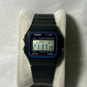 Svart digital klocka från Casio - Säljer en klassisk svart digital klocka från Casio, modell F-91W. Klockan har en rektangulär display med blå och gul text, och funktioner som alarm och kronograf. Armbandet är i svart plast med justerbart spänne. Perfekt för en retro look!