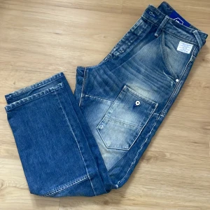 Blå jeans från Jack & Jones - Säljer ett par coola blå jeans från Jack & Jones med en avslappnad look. De har en unik tvättad effekt och praktiska fickor med knappdetaljer. Perfekta för en casual stil.