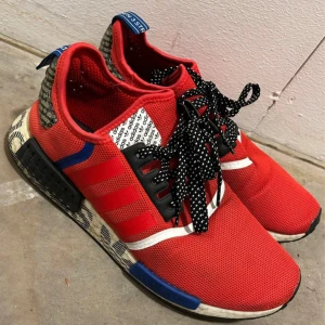 Röda Adidas NMD sneakers - Snygga röda Adidas NMD sneakers med svarta och vita detaljer. Skorna har en stickad ovandel och svarta snören med vita prickar. De har en vit sula med svarta och blå inslag samt en blå hälflik.