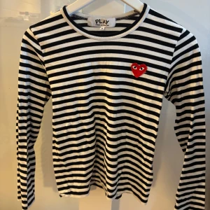 Randig långärmad tröja från Comme des Garçons - Snygg svartvit randig tröja från Comme des Garçons med deras ikoniska röda hjärta på bröstet. Tröjan har långa ärmar och en klassisk rund halsringning. Perfekt för en stilren look.