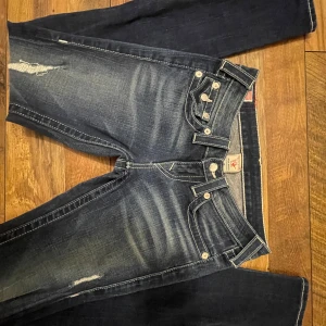 True Religion jeans med slitningar - Snygga mörkblå jeans från True Religion med slitningar och kontrastsömmar. De har en klassisk femficksdesign och knappar med logotyp. Bakfickorna har dekorativa sömmar och knappar. Perfekta för en avslappnad stil.