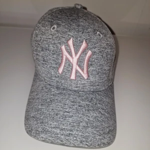 Grå keps med New York logga - Snygg grå keps från New Era med rosa logga. Kepsen har en justerbar rem baktill för perfekt passform. 