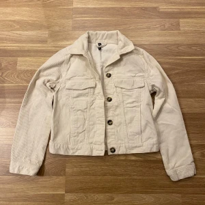 Beige manchesterjacka med knappar - Säljer en snygg beige manchesterjacka med knappar framtill. Jackan har långa ärmar och två bröstfickor med lock. Perfekt för en stilren look!