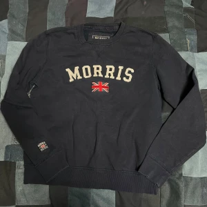 Mörkblå sweatshirt från Morris - Säljer en stilren sweatshirt från Morris med texten 'Morris' och en Union Jack-flagga på bröstet. Tröjan har långa ärmar och ribbade muddar vid ärmslut och nederkant. Perfekt för en avslappnad stil.