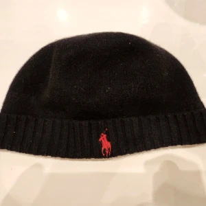 Svart mössa från Ralph Lauren - Snygg svart mössa från Ralph Lauren med röd broderad logga framtill. Den är ribbad vid kanten och perfekt för kyliga dagar. En stilren accessoar som passar till det mesta.