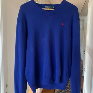 Polo Ralph Lauren pullover v-neck Large herr - Säljer en klassisk blå tröja från Polo Ralph Lauren med v-ringning och det ikoniska röda logotypbroderiet på bröstet. Tröjan är tillverkad i 100% bomull och har långa ärmar. Perfekt för en stilren look.