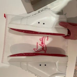 Vita sneakers från Christian Louboutin - Snygga vita sneakers från Christian Louboutin med den ikoniska röda sulan. Skorna har snörning och är tillverkade i skinn, vilket ger en lyxig känsla. Perfekta för dig som vill ha en stilren look med en touch av exklusivitet.