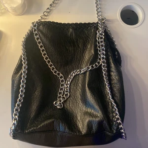 Svart axelväska med kedjedetaljer - Snygg svart axelväska i skinnimitation med coola kedjedetaljer. Perfekt för att ge din outfit en edgy touch. Väskan har en rymlig design. Inga defekter! Nypris:600